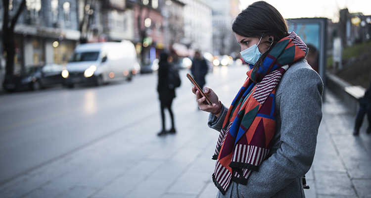 Side view of young woman with face protective mask on the street, she waiting for city transport and looking down, she using smartphone.